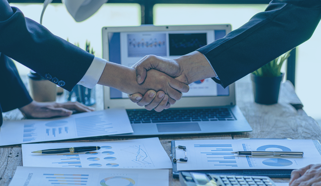 Two professionals shaking hands over a desk covered in charts, documents, and a laptop, symbolizing a successful business agreement.